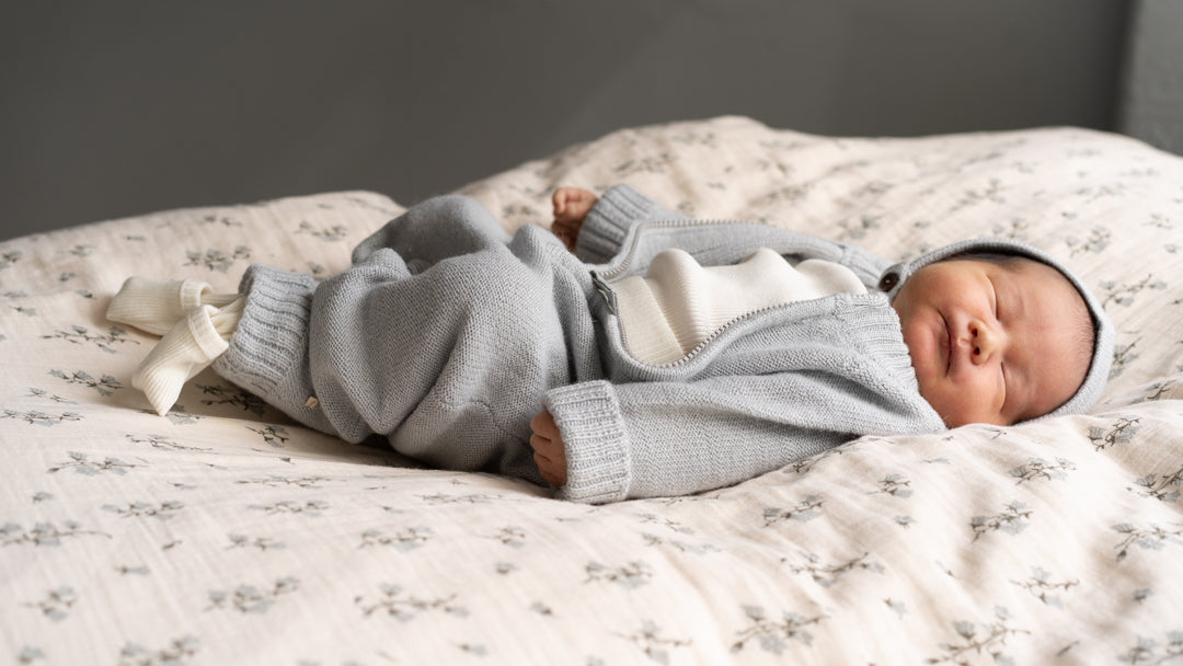A sleeping newborn baby wearing Kys jumsuit in baby-alpaca from minimalisma with a matching bonnet, lying on a floral-patterned cream blanket.